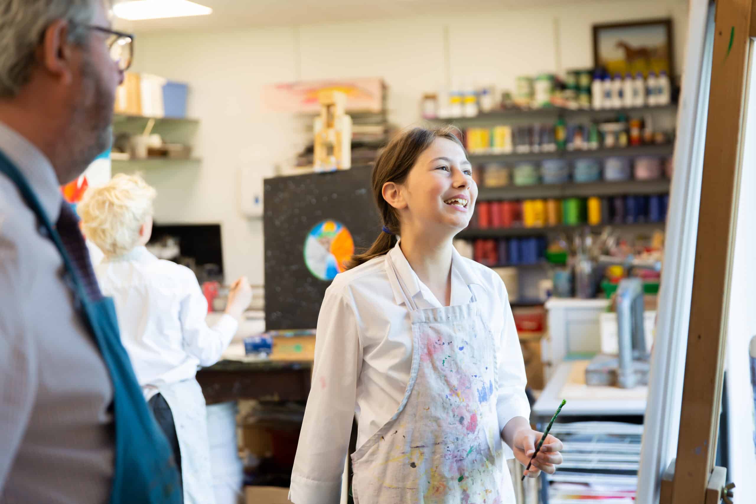 A young girl in a paint-splattered apron smiles while holding a paintbrush in an art classroom. Shelves with various art supplies and colorful paints line the background. An adult in the foreground and another person in the back work on their own art projects, showcasing our About Us passion for creativity.