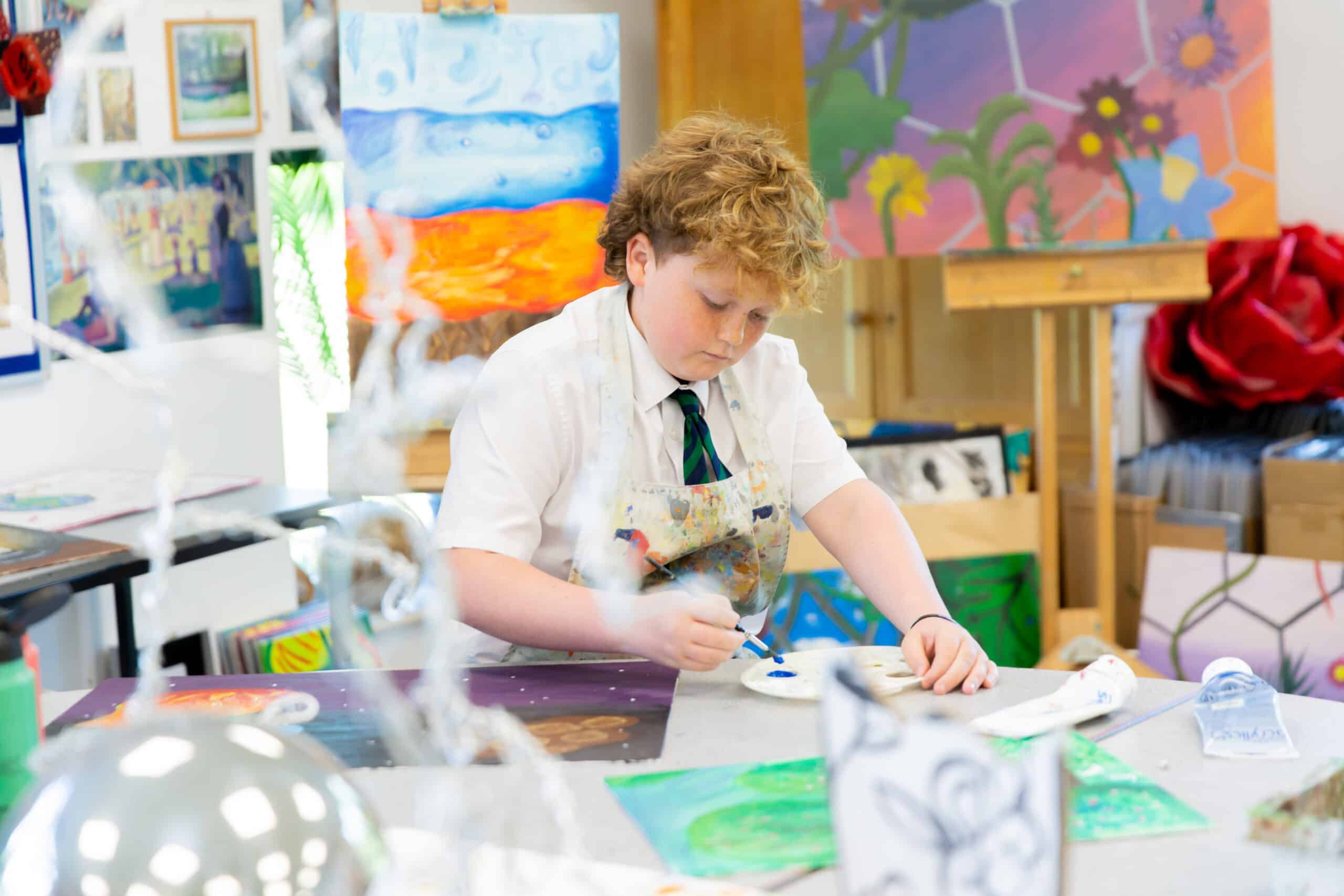 A boy with curly hair is sitting at a table in an art room, painting on a canvas. He wears a white shirt and a patterned tie with a paint-stained apron. The room contains several colorful paintings and art supplies, showcasing an environment that is bright and creative—truly an inspiring art space.