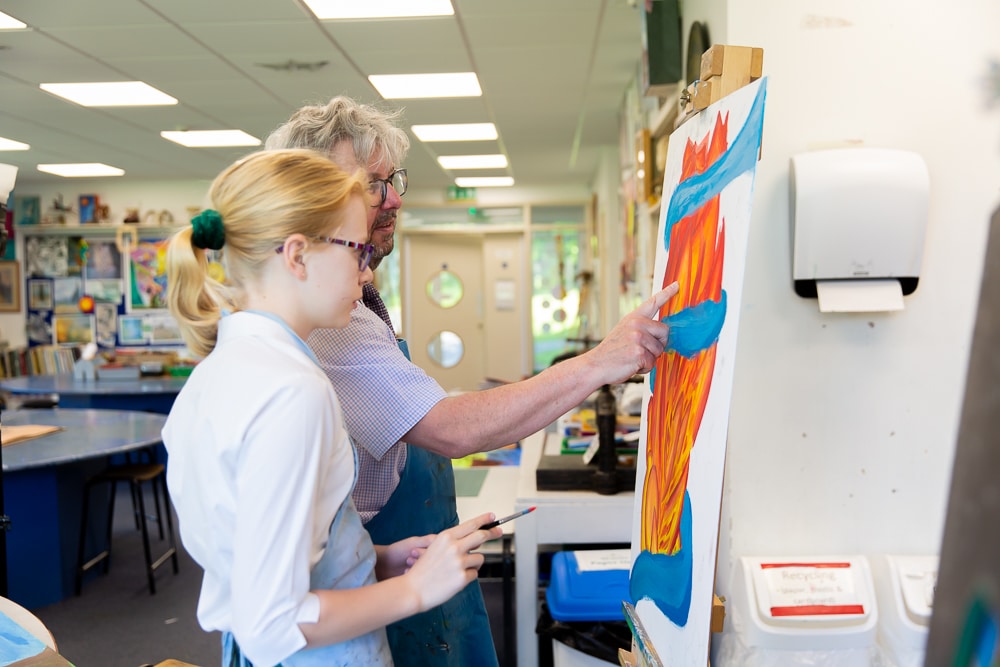 A young person with blond hair, wearing glasses and a blue apron, holds a paintbrush and stands next to an older adult with gray hair and glasses. The adult points to a colorful painting on an easel. They appear to be in an art classroom, surrounded by various supplies.