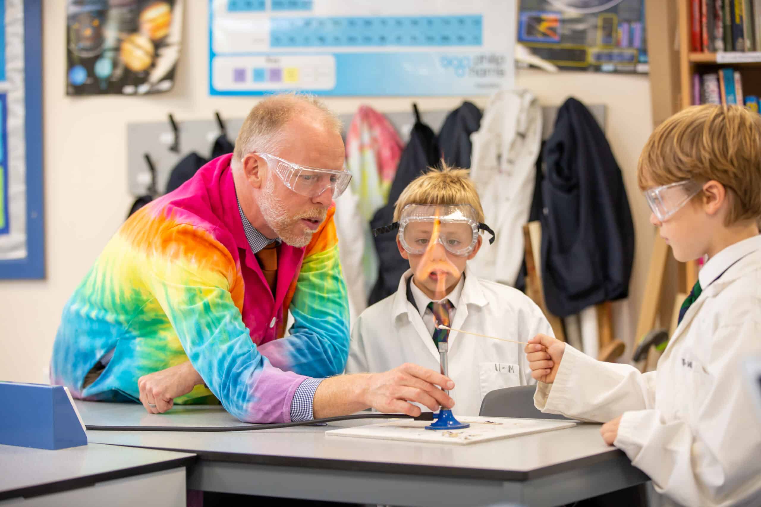 A teacher in a colorful tie-dye lab coat assists two young students, all wearing safety goggles, in a science experiment involving a Bunsen burner in a science classroom. The students and teacher are engaged and focused on the activity.
