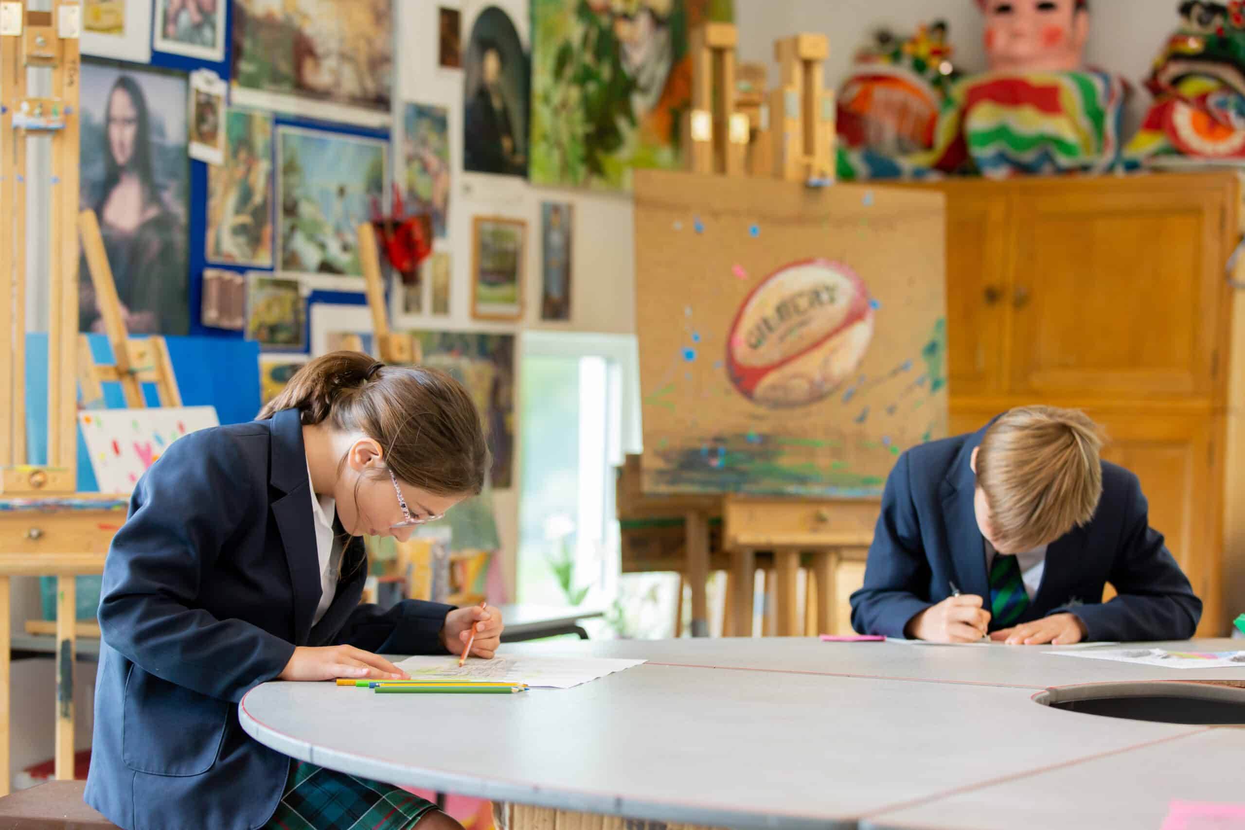 Two students in school uniforms concentrate on their drawings at a table in an art classroom. The room is vibrant, with various artworks, including a Mona Lisa replica, displayed on the walls and a mannequin adorned with colorful fabric in the background, epitomizing their immersion in art.