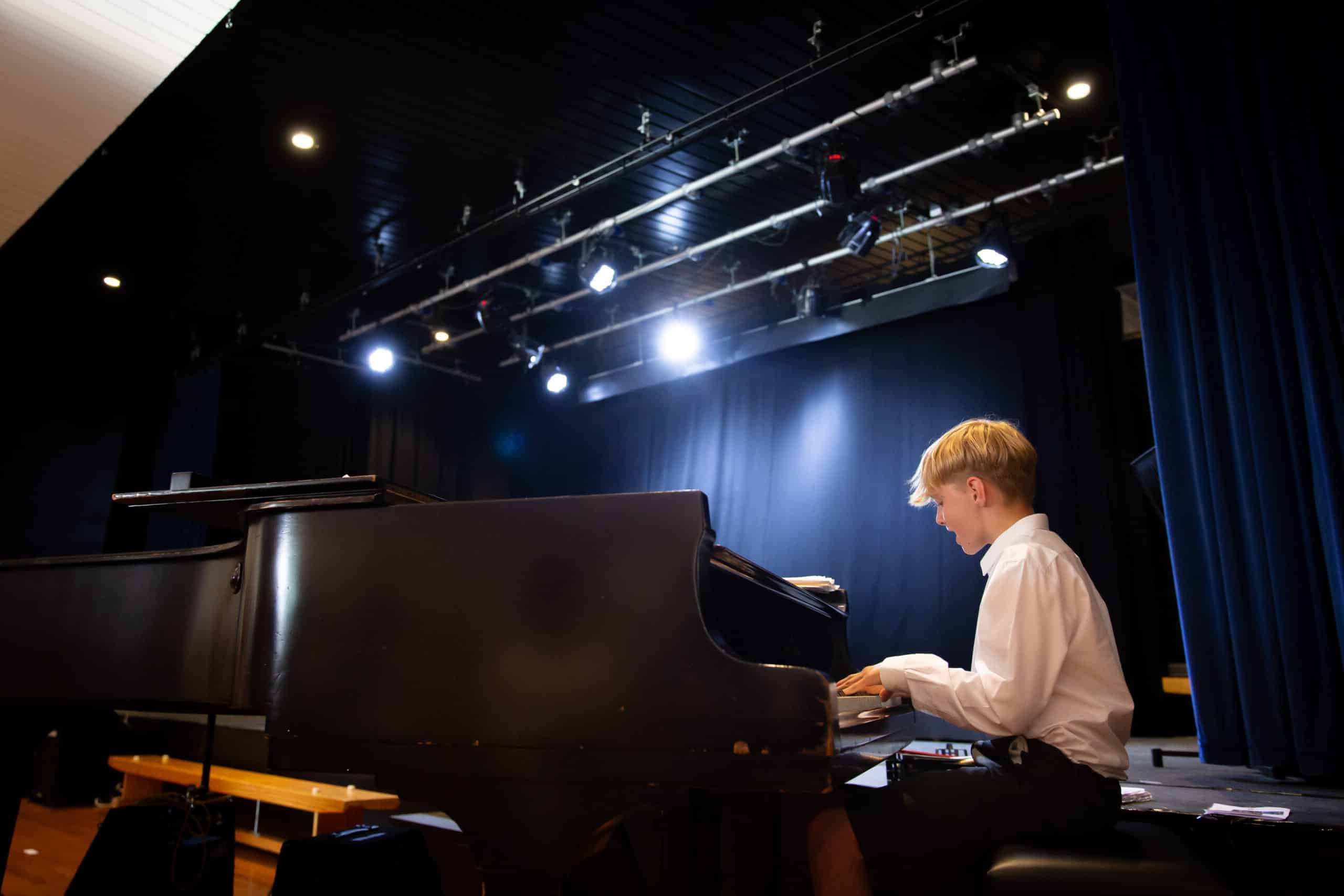 A young boy in a white dress shirt and black pants sits on a piano bench, playing the grand piano on a stage under bright stage lights. With dark curtains and wooden benches in the background, the scene is filled with the enchanting essence of music.